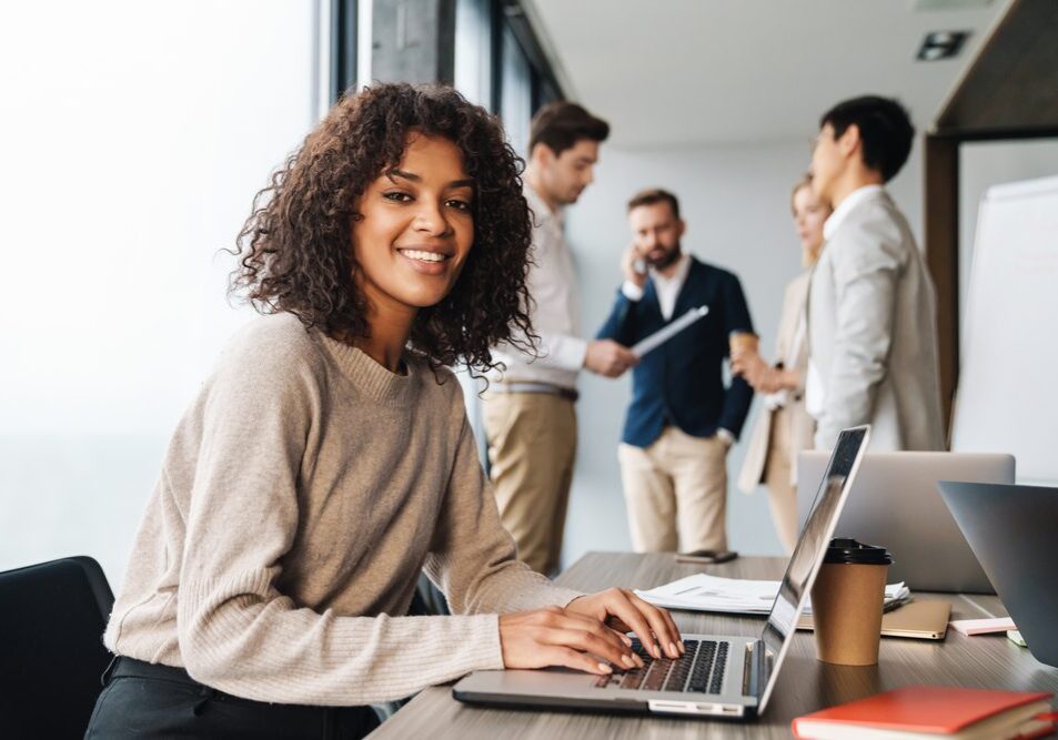 Attractive african young confident businesswoman sitting at the office table with group of colleagues in the background, working on laptop computer